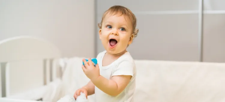 Wesołe dziecko z szeroko otwartymi ustami i oczami trzyma w rączce niebieski smoczek, stojąc w łóżeczku