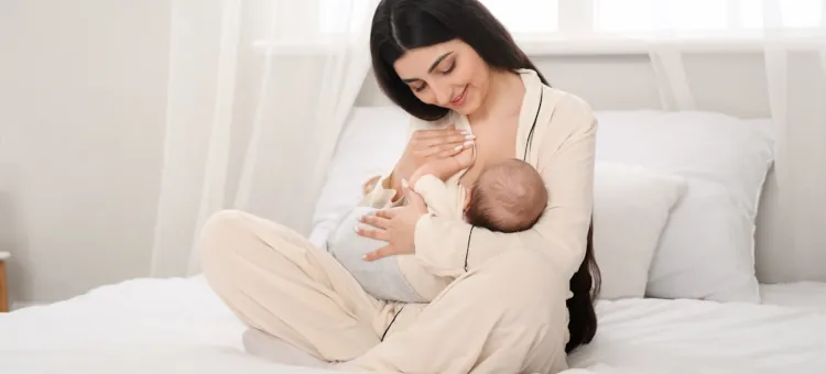 Młoda radosna kobieta z czułością karmiąca piersia noworodka, siedząc na białej pościeli