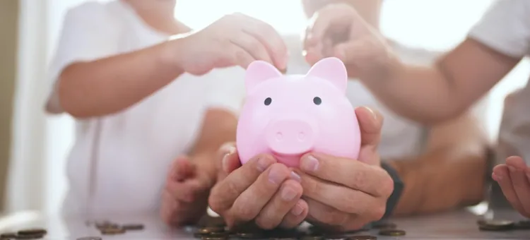 Dziecięce i dorosłe ręce wrzucające bilon do różowego pojemnika na oszczędności w kształcie świnki skarbonki