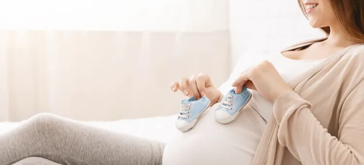 Uśmiechnięta kobieta w zaawansowanej ciąży prezentuje jasnoniebieskie buciki dziecięce na swoim brzuchu