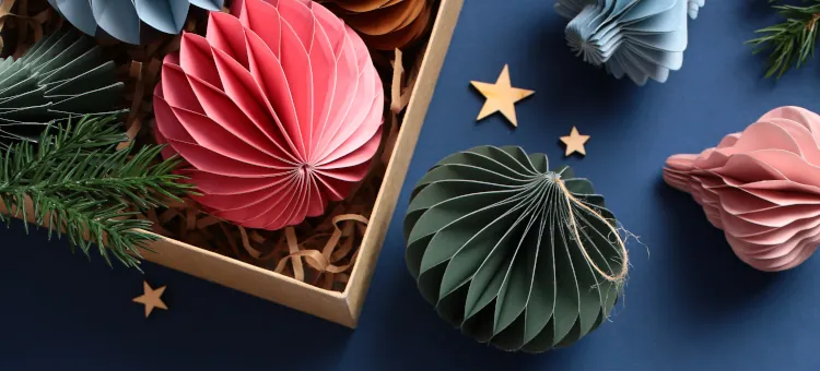 Ozdoby świąteczne, które zrobisz samodzielnie – inspiracje na wyjątkowe dekoracje Różnorodne ozdoby z papieru w pastelowych kolorach, ułożone w pudełku i poza nim na ciemnogranatowym tle, z gałązką drzewa iglastego