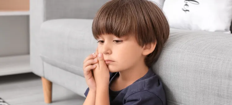 Wyrywanie zęba mlecznego u dziecka – wskazania i najczęstsze powody Dziecko z brązowymi włosami ma zamyślony wyraz twarzy, siedząc obok szarej kanapy, trzyma się za policzek, co wskazuje na ból zęba