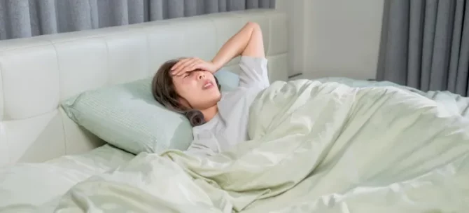 Młoda kobieta z gorączką leży w pościeli, dotykając czoła