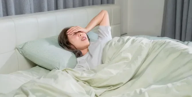 Młoda kobieta z gorączką leży w pościeli, dotykając czoła