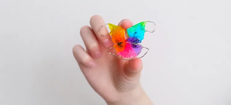 FAQ Dłoń dziecka prezentująca barwny aparat ortodontyczny do korekcji zgryzu