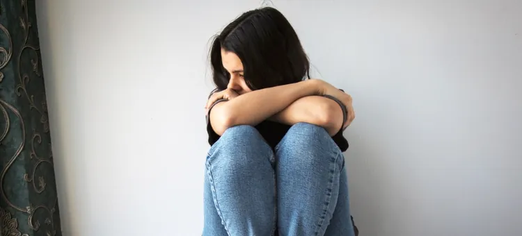 Najczęstsze przyczyny poronienia zatrzymanego Smutna kobieta siedząca, zamyślona, oparta o białą ścianę, z wiszącą z lewej strony ciemną zasłoną