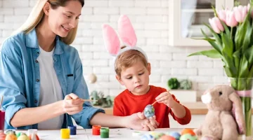 Chłopiec w opasce z uszami królika i radosna kobieta ozdabiają razem świąteczne jajka na jasnym blacie w kuchni