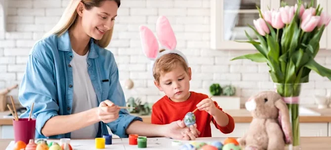 Chłopiec w opasce z uszami królika i radosna kobieta ozdabiają razem świąteczne jajka na jasnym blacie w kuchni