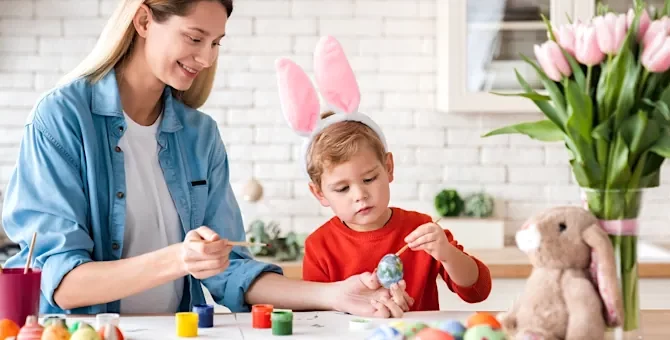 Chłopiec w opasce z uszami królika i radosna kobieta ozdabiają razem świąteczne jajka na jasnym blacie w kuchni