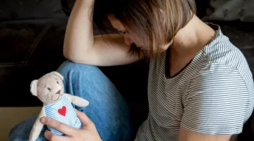 Zamyślona postać kobieca w bluzce w paski, oparta o ciemny narożnik, trzyma niedźwiadka- zabawkę