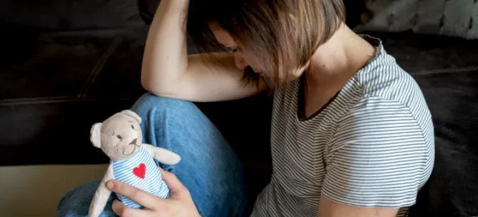 Zamyślona postać kobieca w bluzce w paski, oparta o ciemny narożnik, trzyma niedźwiadka- zabawkę