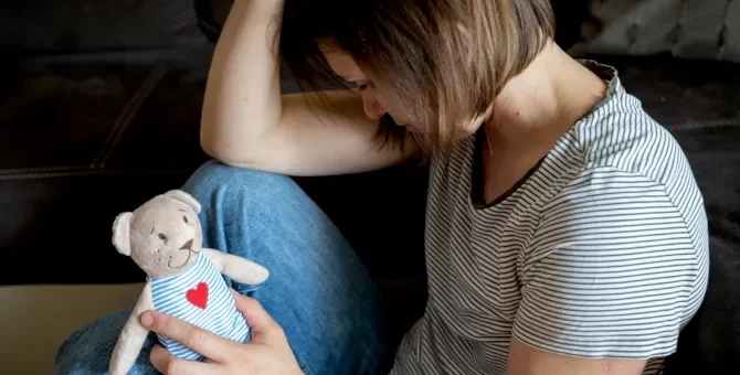 Zamyślona postać kobieca w bluzce w paski, oparta o ciemny narożnik, trzyma niedźwiadka- zabawkę