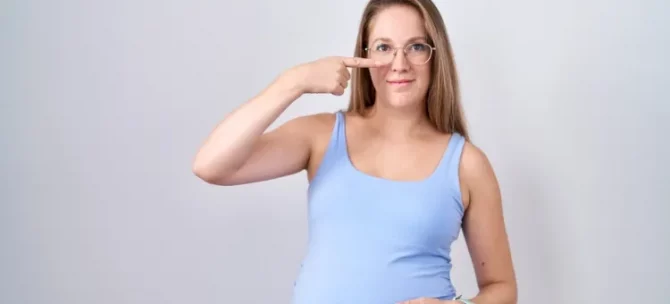 Ciężarna, uśmiechnięta pani w okularach na nosie, pokazuje swój nos