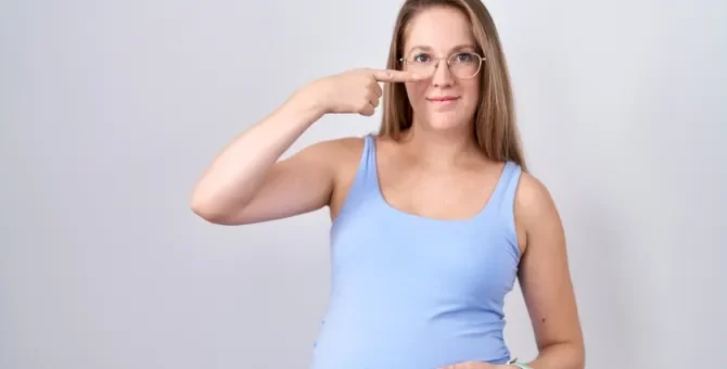 Ciężarna, uśmiechnięta pani w okularach na nosie, pokazuje swój nos