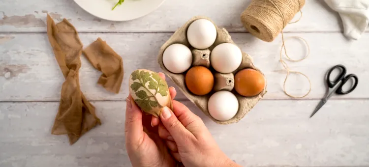 Dłoń ozdabiająca skorupkę liściem przed barwną kąpielą jajek w tekturowym pudełku na bielonych deskach