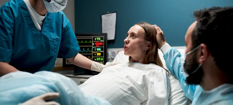 Kobieta w jasnym stroju na łóżku w trakcie porodu ze wsparciem medyków i bliskiej osoby