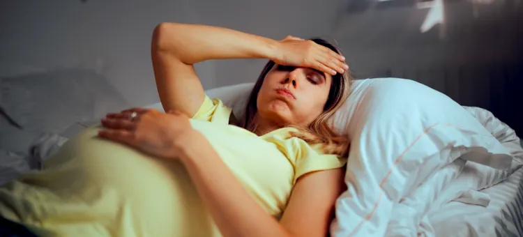 Katar w ciąży a przeziębienie – kiedy katar jest objawem infekcji wirusowej lub bakteryjnej? Przyszła mama odczuwająca ból głowy spoczywa na białej pościeli, przykładając rękę do czoła