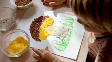 Małe dziecięce dłonie w trakcie zajęć sensorycznych formują obraz kaszą na jasnym drewnianym blacie