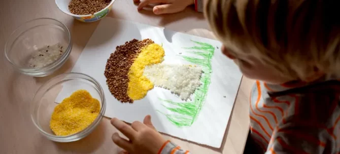 Małe dziecięce dłonie w trakcie zajęć sensorycznych formują obraz kaszą na jasnym drewnianym blacie