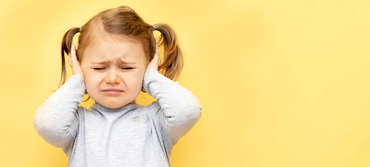 Mała dziewczynka ze zmarszczoną twarzą zasłania uszy przed nieprzyjemnymi dźwiękami, na gładkim żółtym tle