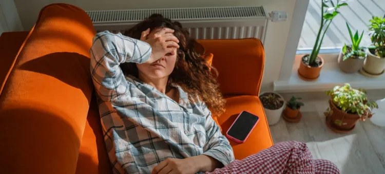 Młoda kobieta zmaga się z wyczerpaniem, osłaniając twarz, na jasnej pomarańczowej sofie