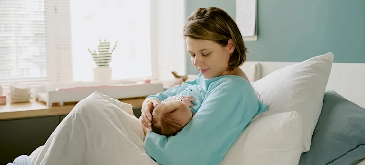 Matka z czułością karmi maleństwo piersią w łóżku przy jasnoniebieskiej ścianie