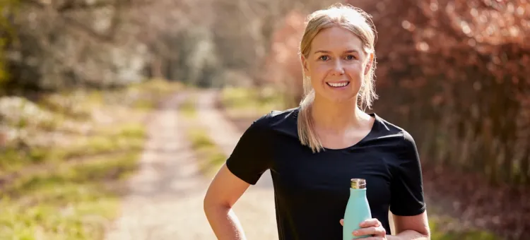 Energetyczna blondynka w czarnym t-shircie trzyma turkusową butelkę nawadniającą na tle słonecznej, brązowo-zielonej leśnej ścieżki
