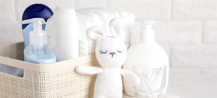 Uporządkowany zestaw akcesoriów dziecięcych z pieluszkami i miękkim królikiem w wiklinowym pojemniku na jasnym, białym tle ściany