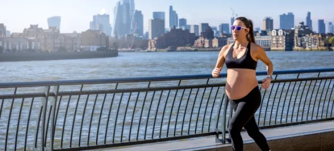 Aktywnie ćwicząca przyszła mama pokonuje trasę po jasnej betonowej nawierzchni, z rzeką o granatowej wodzie i miejskim pejzażem w słonecznym tle