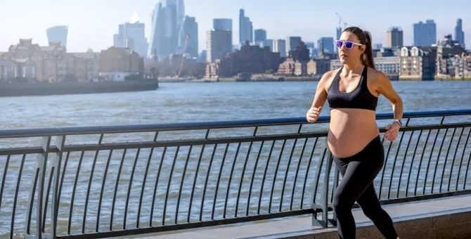 Aktywnie ćwicząca przyszła mama pokonuje trasę po jasnej betonowej nawierzchni, z rzeką o granatowej wodzie i miejskim pejzażem w słonecznym tle