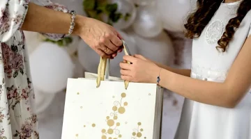 Kobieta przekazuje elegancki upominek w ozdobnej torebce dziecku w białej sukience komunijnej, celebrując Komunię Świętą, zbliżenie na ręce i prezent