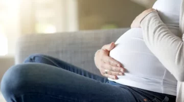 Przyszła mama w białej koszulce czule obejmuje dłońmi duży brzuszek, odpoczywając na jasnoszarej sofie