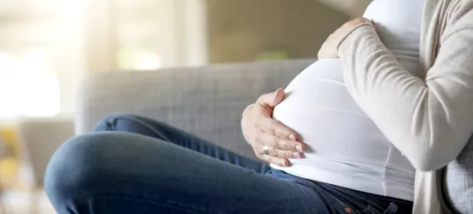 Przyszła mama w białej koszulce czule obejmuje dłońmi duży brzuszek, odpoczywając na jasnoszarej sofie