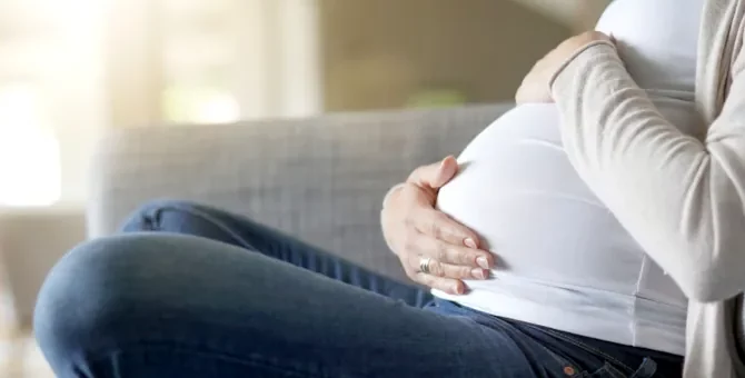 Przyszła mama w białej koszulce czule obejmuje dłońmi duży brzuszek, odpoczywając na jasnoszarej sofie