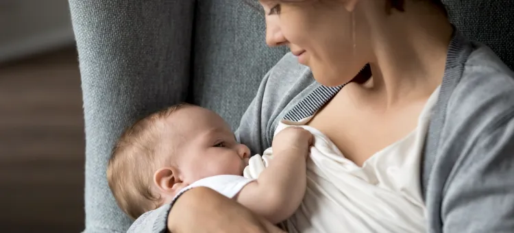 Co robić, aby uniknąć zachorowania na zapalenie oskrzelików u dzieci? Szczęśliwa mama trzyma w ramionach maluszka podczas naturalnego karmienia, siedząc w szarym fotelu