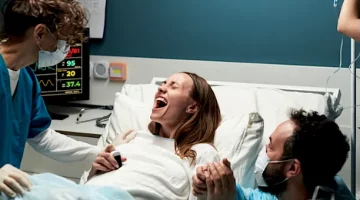 Pacjentka na łóżku porodowym głośno krzyczy podczas skurczu, mocno ściskając dłoń ukochanego na tle ciemnoturkusowej ściany