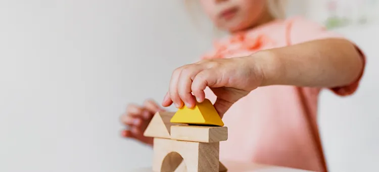 Adaptacja dziecka w przedszkolu może potrwać dłużej, niż myślisz Mała dziewczynka w różowym ubranku precyzyjnie kładzie żółty trójkątny element na wieży z ekologicznego surowca na jasnym tle, zbliżenie na ręce