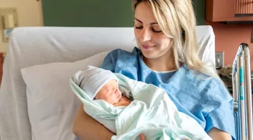 Uśmiechnięta kobieta w błękitnej koszuli opiekuje się małym dzieckiem w jasnym otoczeniu medycznym na tle zielonej ściany