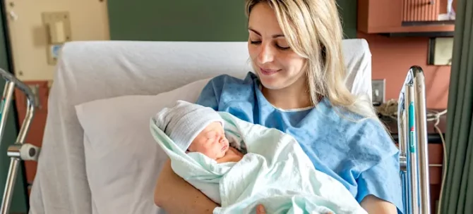 Uśmiechnięta kobieta w błękitnej koszuli opiekuje się małym dzieckiem w jasnym otoczeniu medycznym na tle zielonej ściany