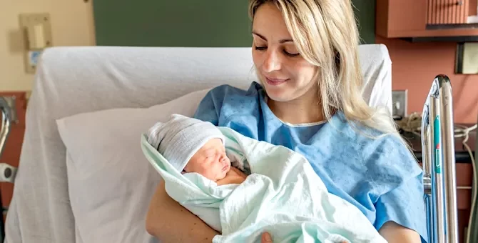 Uśmiechnięta kobieta w błękitnej koszuli opiekuje się małym dzieckiem w jasnym otoczeniu medycznym na tle zielonej ściany