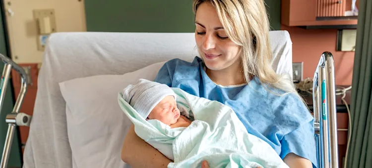 Uśmiechnięta kobieta w błękitnej koszuli opiekuje się małym dzieckiem w jasnym otoczeniu medycznym na tle zielonej ściany