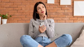 Zamyślona dziewczyna trzyma telefon komórkowy, siedząc na szarym kanapie na tle pomarańczowej ściany z cegły