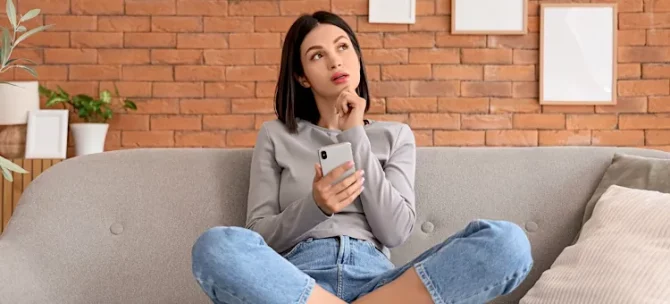 Zamyślona dziewczyna trzyma telefon komórkowy, siedząc na szarym kanapie na tle pomarańczowej ściany z cegły