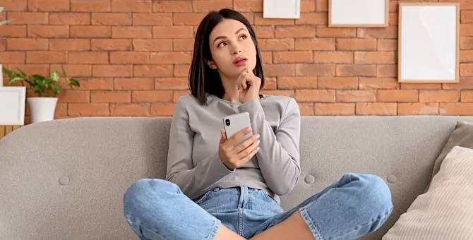 Zamyślona dziewczyna trzyma telefon komórkowy, siedząc na szarym kanapie na tle pomarańczowej ściany z cegły