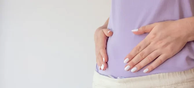 Dłonie z jasnym manicure ułożone na jasnofioletowym stroju okrywającym brzuch ciążowy, na jasnym tle