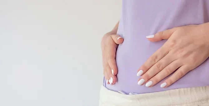 Dłonie z jasnym manicure ułożone na jasnofioletowym stroju okrywającym brzuch ciążowy, na jasnym tle