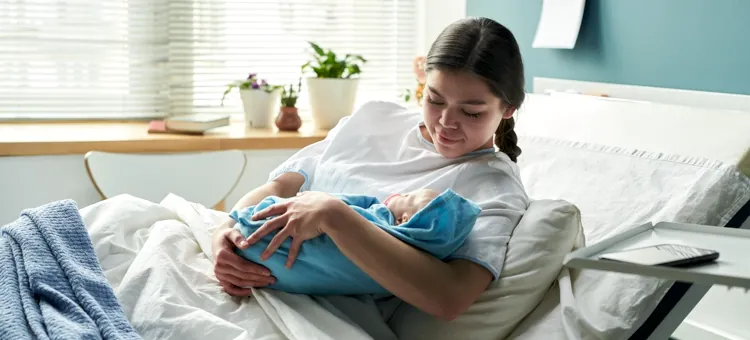 Dlaczego dochodzi do syndromu drugiej doby? Pacjentka trzyma w ramionach owinięte niebieskim kocykiem dziecko, leżąc w szpitalnym łóżku na tle jasnoniebieskiej ściany