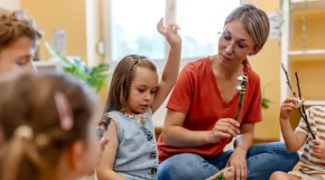 Kobieta w czerwonej koszulce prowadzi zajęcia edukacyjne z grupą przedszkolaków w jasnym pomieszczeniu