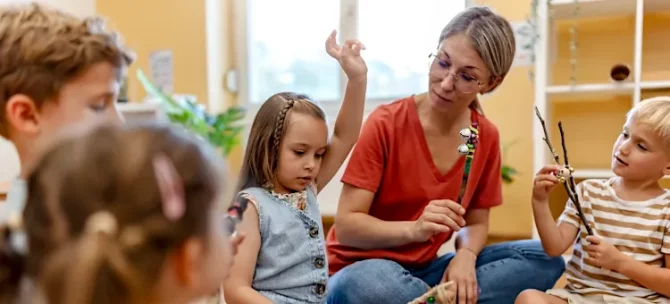 Kobieta w czerwonej koszulce prowadzi zajęcia edukacyjne z grupą przedszkolaków w jasnym pomieszczeniu
