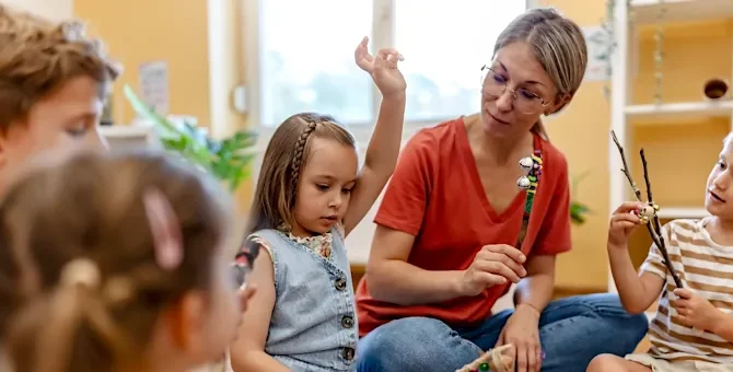 Kobieta w czerwonej koszulce prowadzi zajęcia edukacyjne z grupą przedszkolaków w jasnym pomieszczeniu
